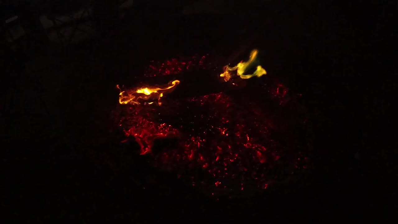 At 0:01, the fire remains low and dark, with red coals spread across the lower frame and two small yellow flame points, no visible people.