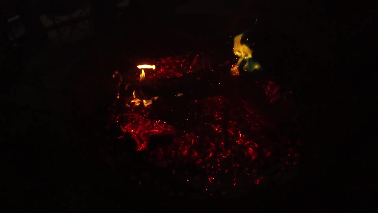 At 0:03, the coals sit low in the dark frame while small flames appear along the left edge and upper area, no visible people.