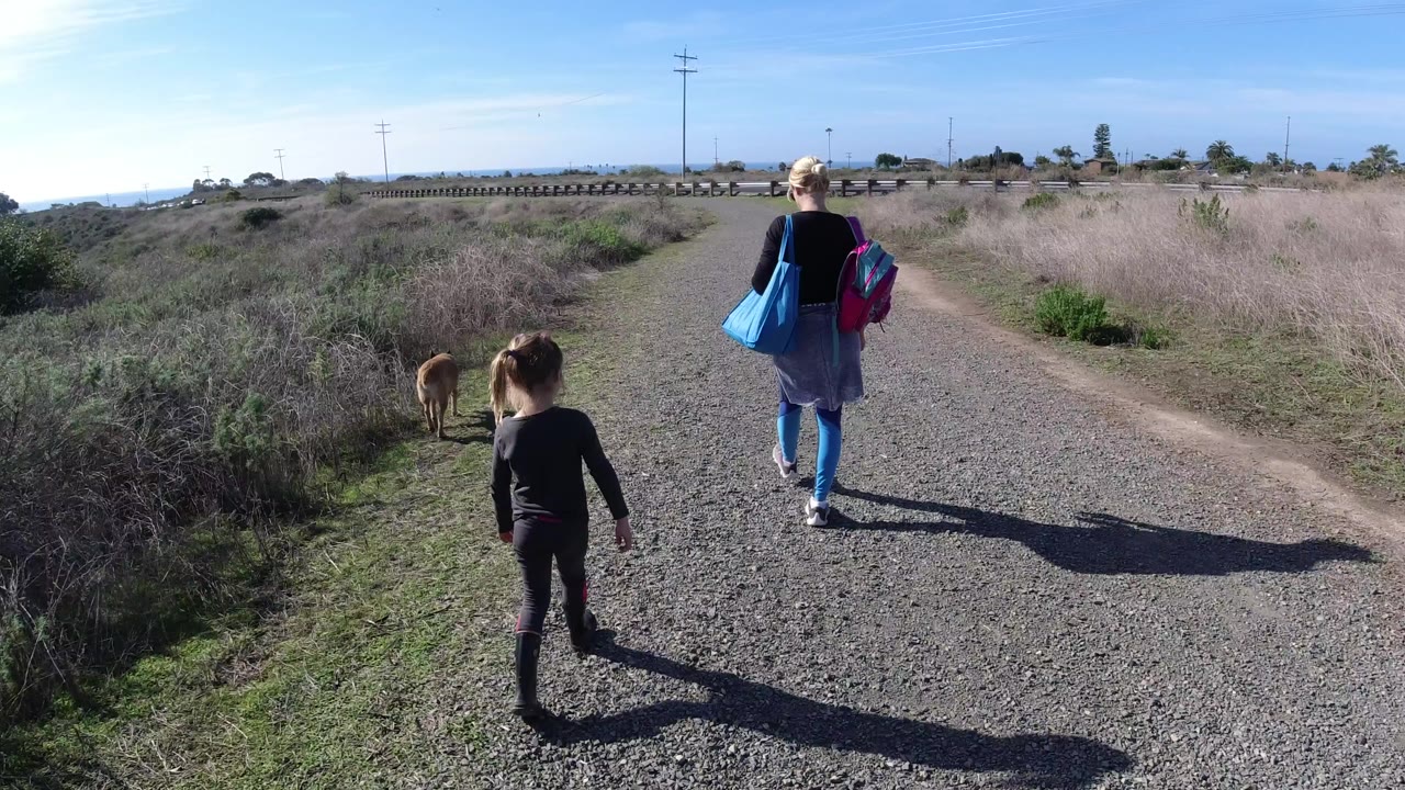 In San Clemente, California, a woman with bright bags walks ahead of a child and dog on a gravel road bordered by dry brush and low greenery.