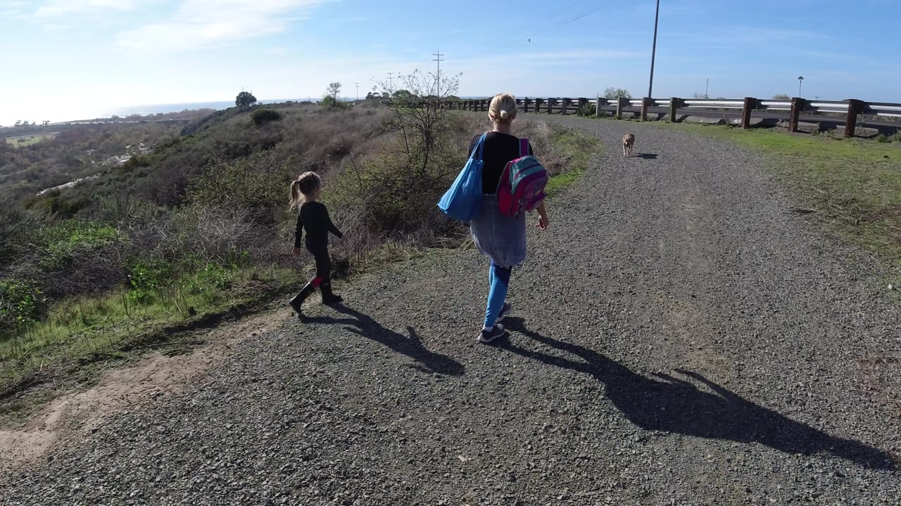 Two people continue down the gravel trail in San Clemente, California, while the dog stays ahead and the bright sun creates dark shadows stretching across the path.