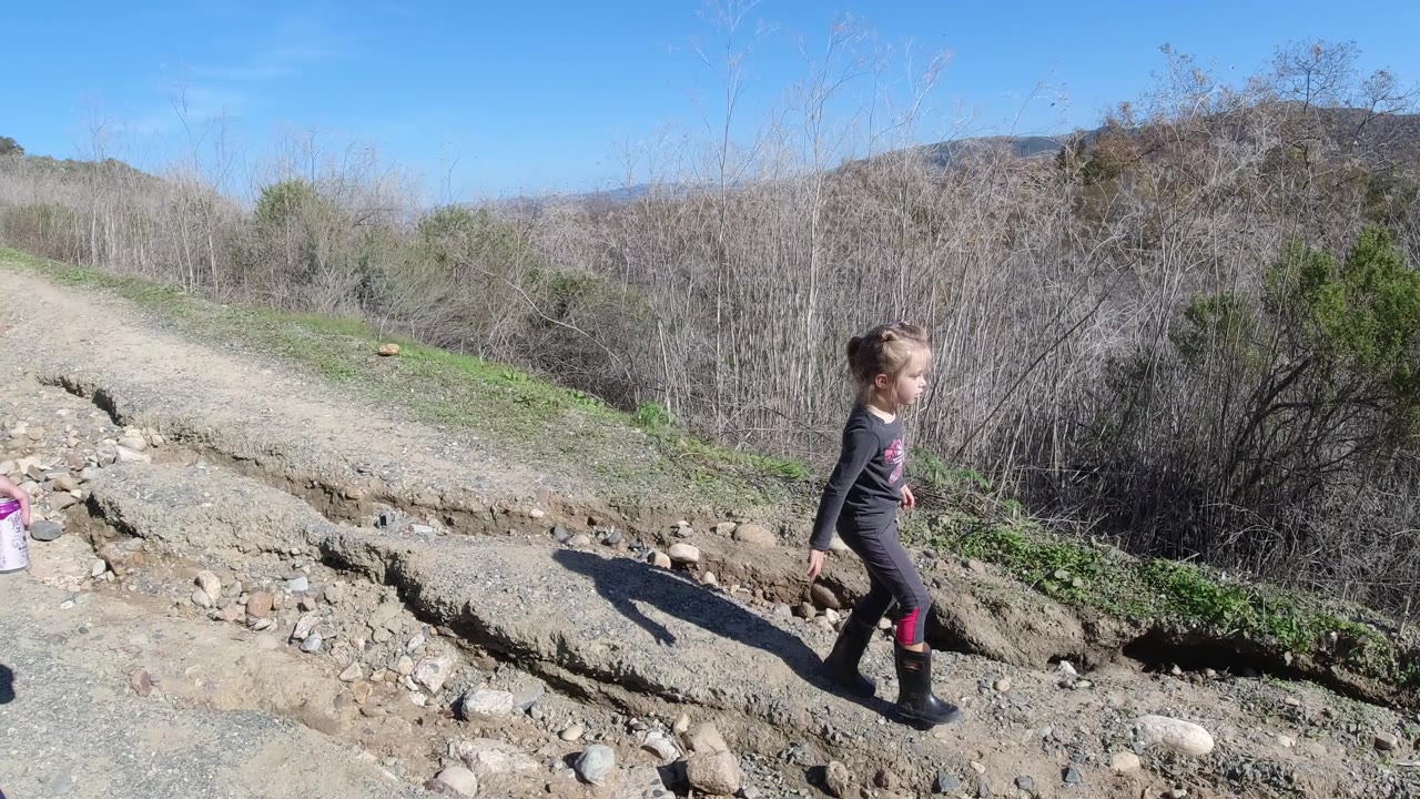 A child walks along a rocky dirt path in San Clemente, California, with eroded ground, low brush, distant hills, and bright blue sky behind her.