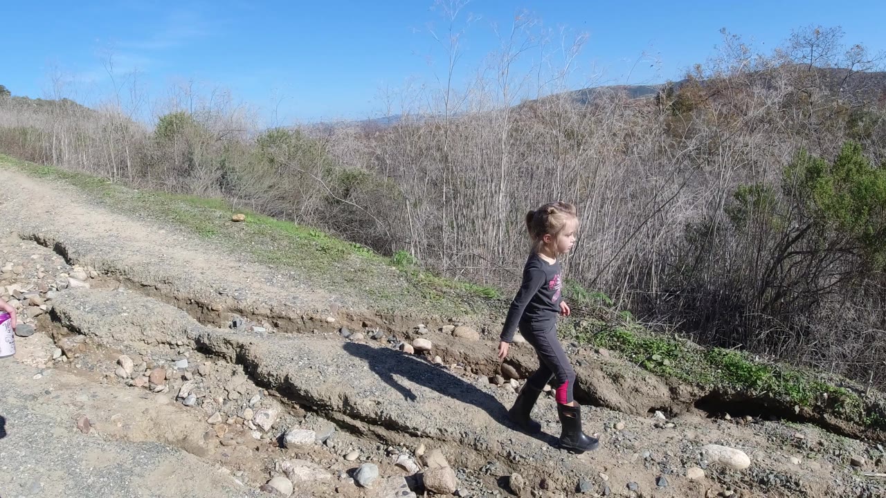 Child walking beside an eroded dirt trail with brush and hills.