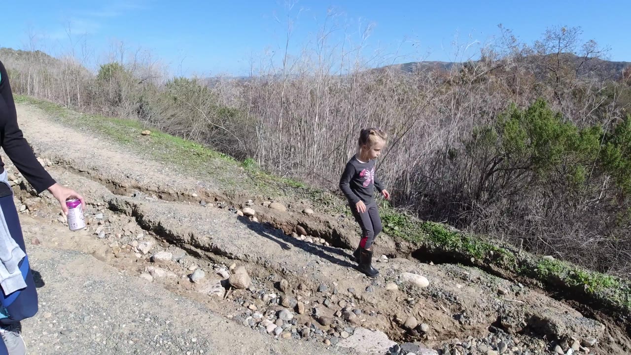 An adult woman is partly visible at the right edge while the child walks on the San Clemente dirt trail beside brush, rocks, and low green growth.