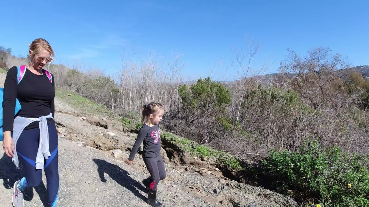 At the start of the San Clemente clip, a woman and young child walk along a dusty hillside trail with brush and low hills behind them.