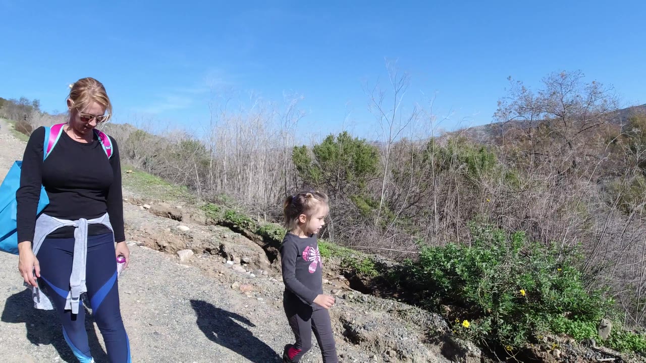 At 0:02, the woman with a bright backpack and the child pass through the frame with open sky, shrubs, and a low hillside in the background.