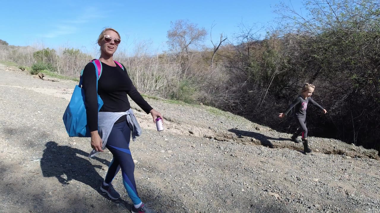 A smiling woman with a large blue bag walks past the camera on a rocky San Clemente path while a young child moves near the dark brush at right.