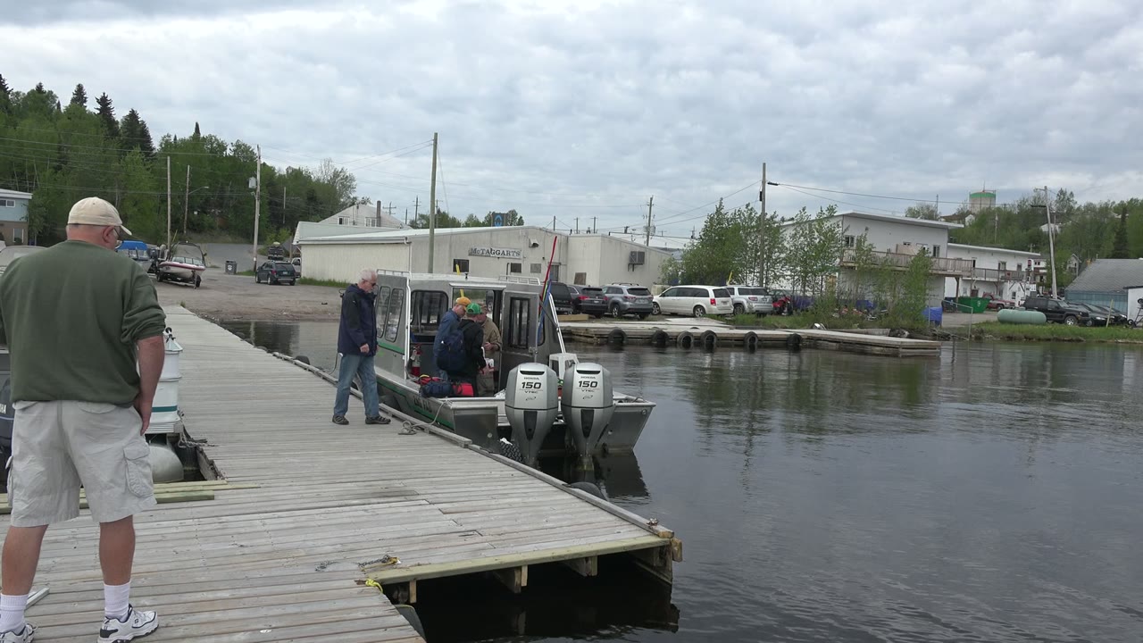 Adults stand on a dock beside a small motorboat on calm water.