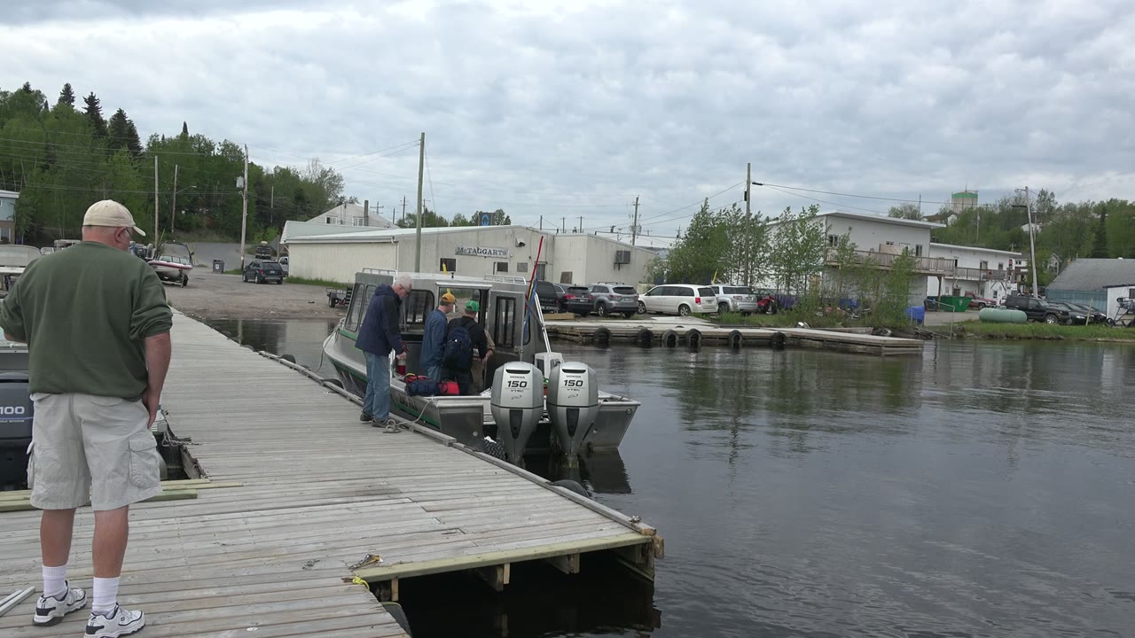 A man in shorts stands in the foreground on a Red Lake dock as other adults work around the stern of a small motorboat beside reflective water.