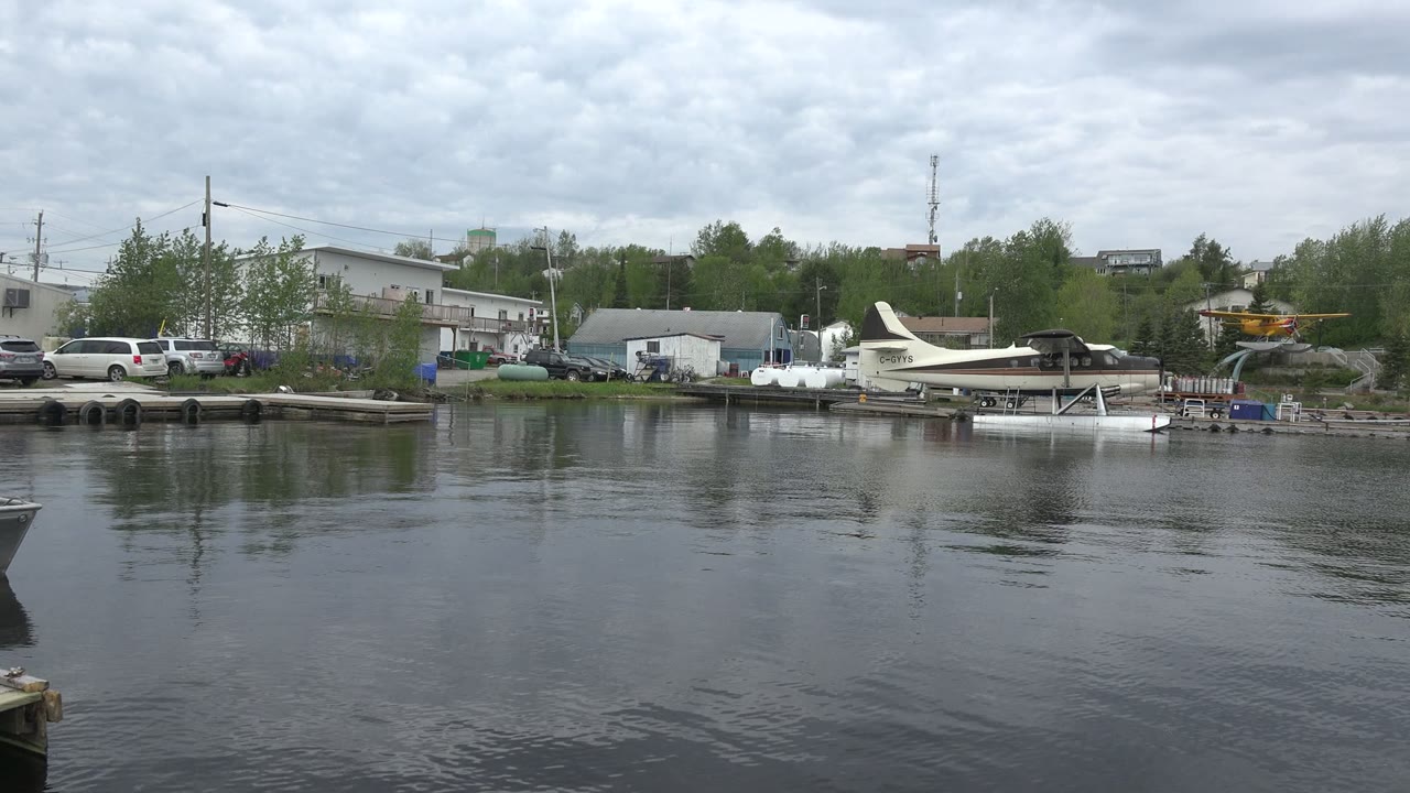 Wide lake view with shoreline buildings, docks, cars, and seaplanes under gray clouds.