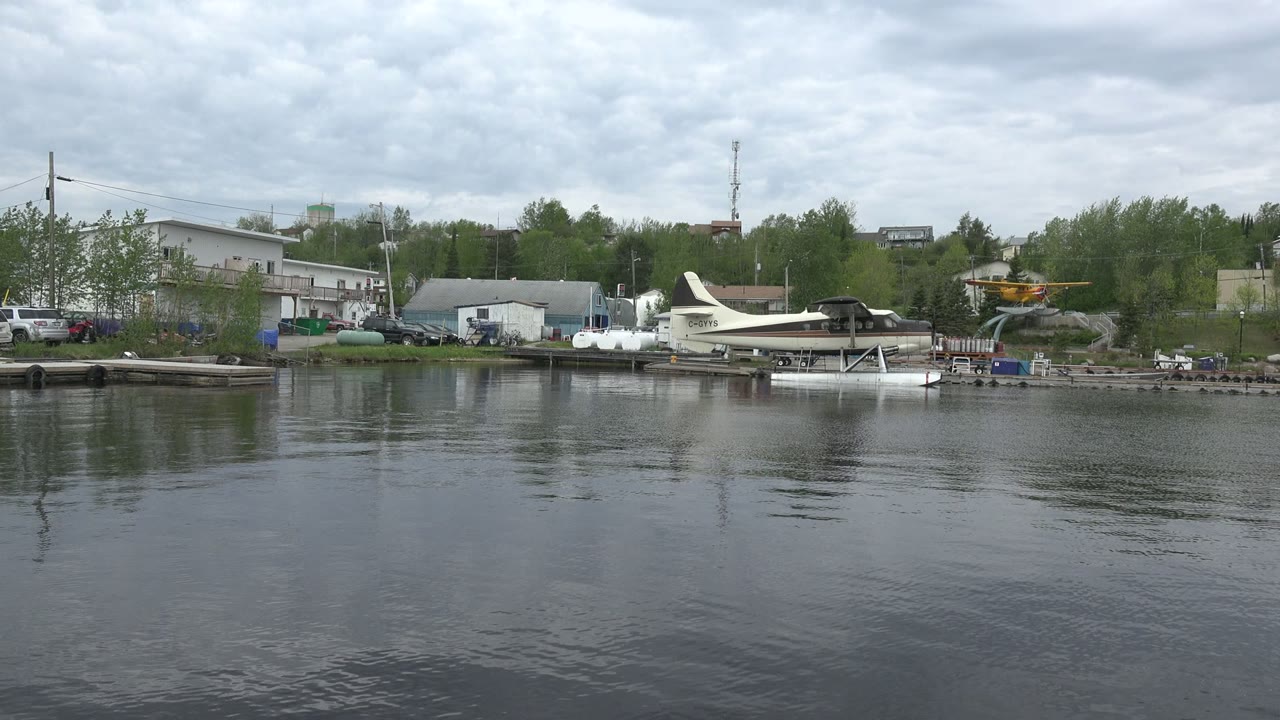 Red Lake, Canada shoreline view with a white seaplane on floats near docks, parked cars by low buildings, green trees, and a yellow aircraft entering at the right edge, with no visible people.