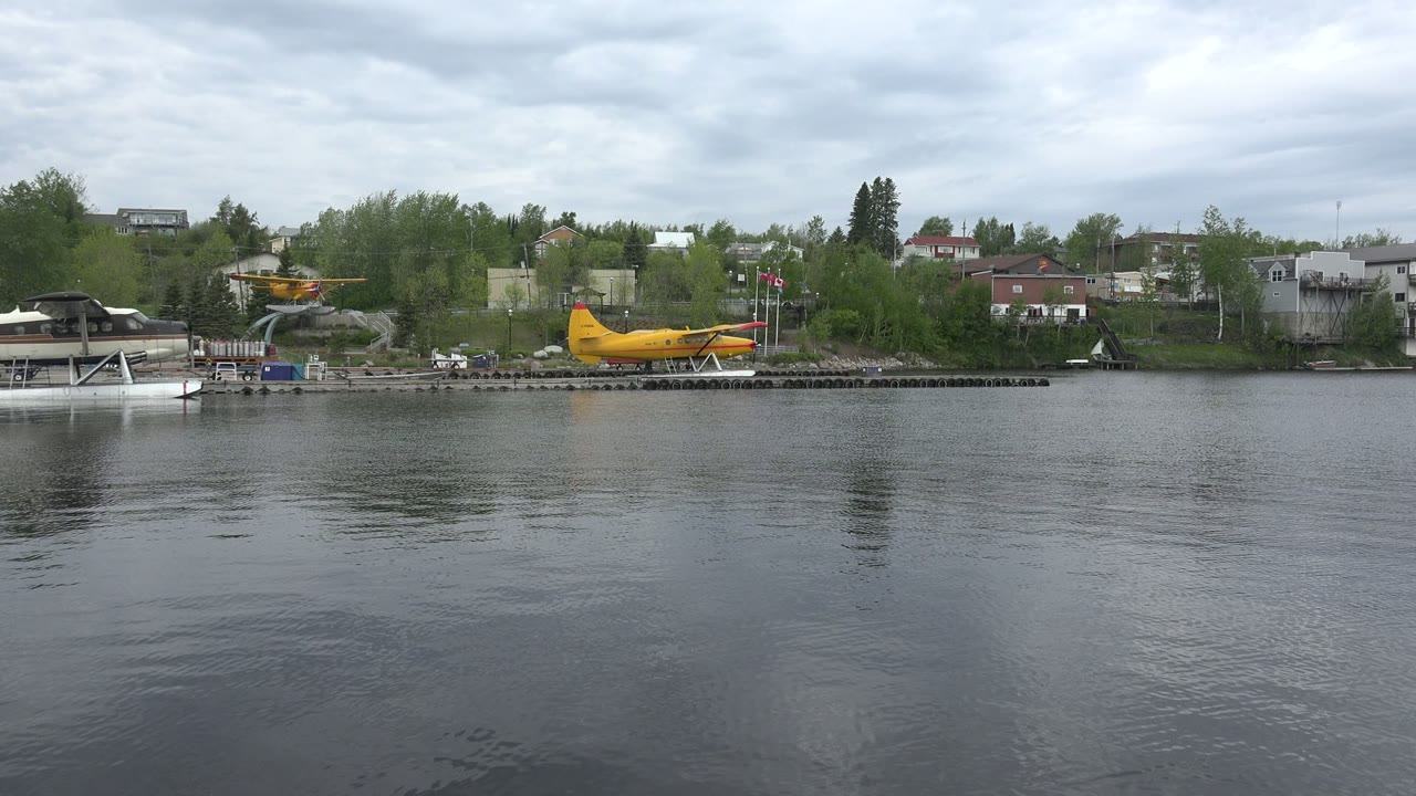 At 0:01, a yellow seaplane sits near the center of the Red Lake, Canada shoreline, with calm gray water, trees, buildings, and no visible people.