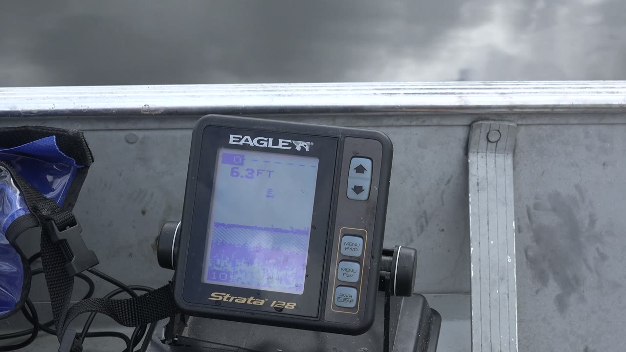 At 0:00, a black Eagle Strata 128 display shows a depth reading beside a metal boat side on Red Lake, Canada, with grey water behind it and no visible people.