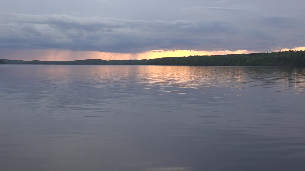 Opening frame of Red Lake, Canada with blue-gray lake water in the foreground, a dark tree line on the horizon, warm sunset light beneath thick clouds, and no visible people.