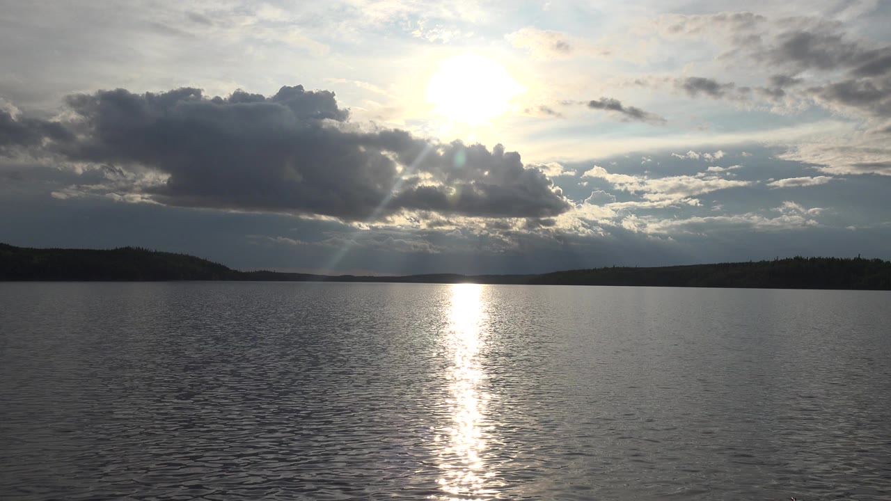 At 0:00 in Red Lake, Canada, the sun glows behind broken clouds over rippled lake water, with a vertical reflection and no visible people.