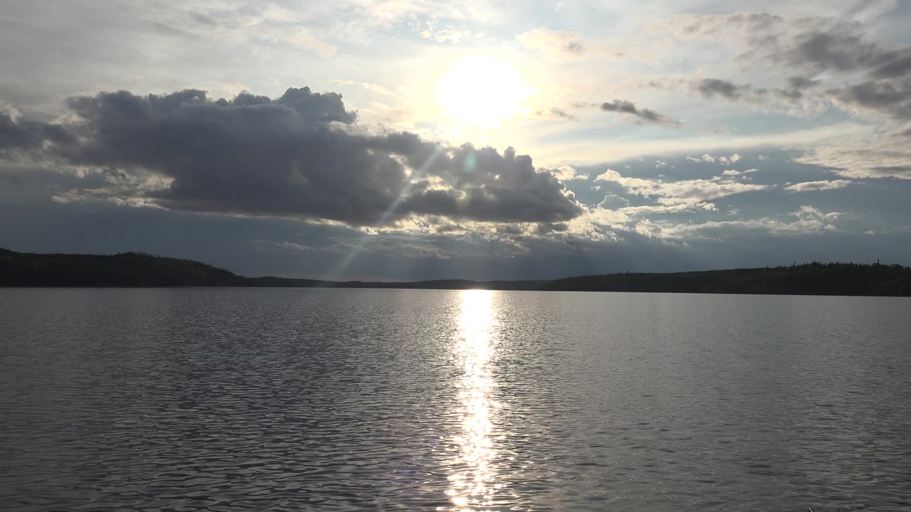 At 0:01 in Red Lake, Canada, a large dark cloud sits below the high sun while the lake reflects a narrow bright strip, with no visible people.