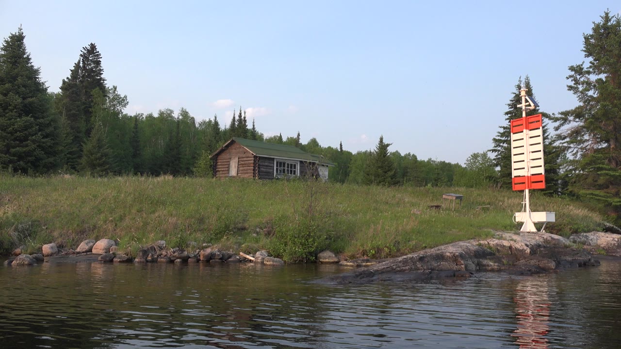 At 0:00 in Red Lake, Canada, rippled water fills the foreground below a rocky grassy shore with a small house, tall trees, blue sky, and a red and white marker at right, with no visible people.