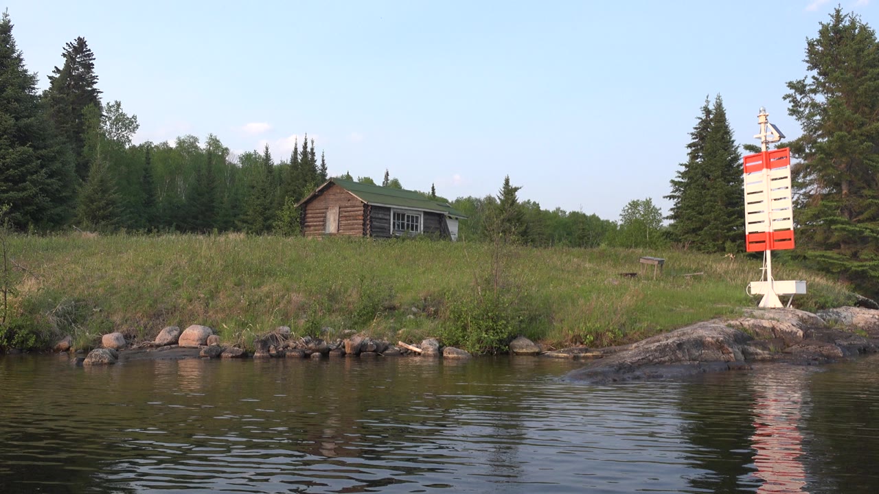 Opening frame of Red Lake, Canada with rippled water in front, a grassy rocky bank, a small wooden cabin left of center, a red and white shoreline marker to the right, and no visible people.