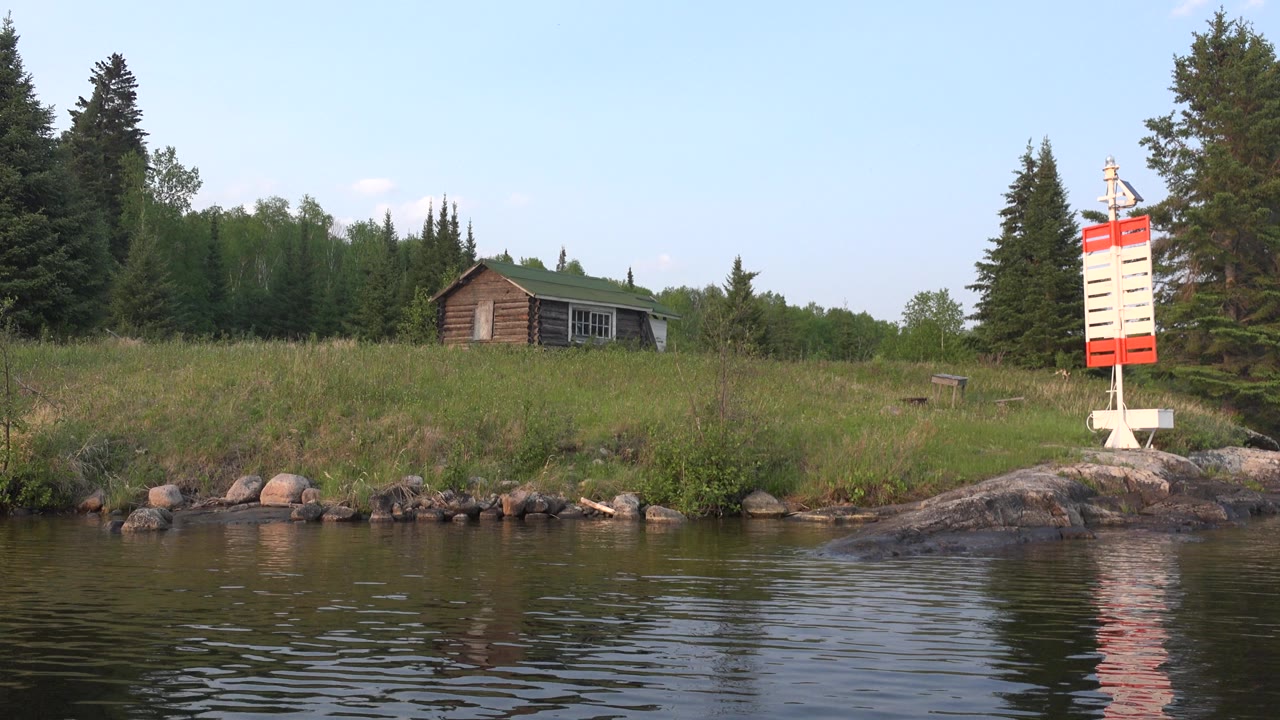 At 0:01 in Red Lake, Canada, the water ripples shift slightly below the same grassy bank, cabin, tree line, and red and white marker, with no visible people.
