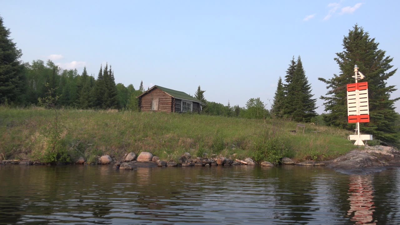 Small lakeside house, trees, shoreline rocks, water, and red and white marker