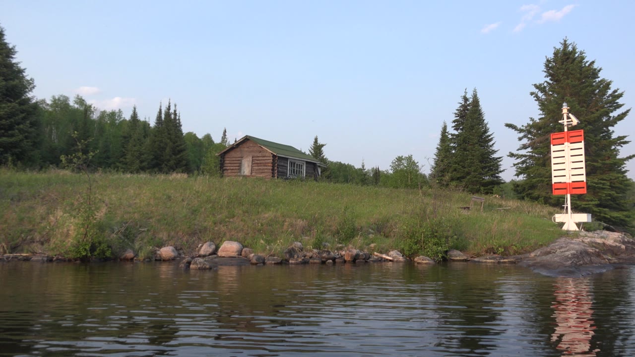 Red Lake, Canada shoreline frame showing the house slightly left of center, dark evergreens behind it, rippled foreground water, stone bank, and the red and white marker at the right edge, with no visible people.