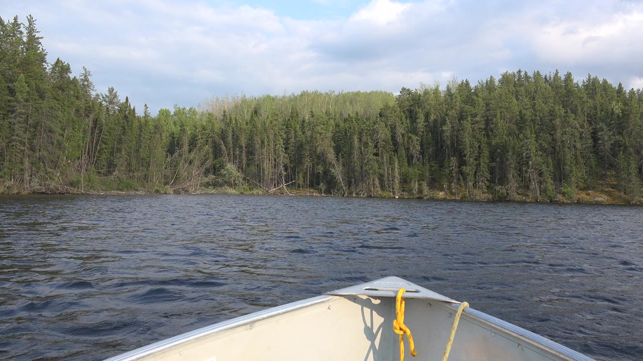 Canoe bow with a yellow rope points across rippled Red Lake water toward a dense green forest shoreline under a cloudy sky, with no visible people.
