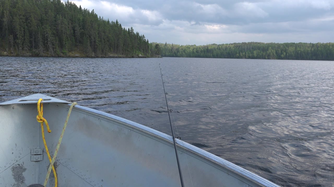 Boat bow and fishing rod on a rippled lake under gray clouds.