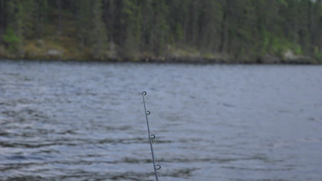 At 0:00 in Red Lake, Canada, a fishing rod tip rises from the lower frame over blue-gray rippled water with a soft forest shoreline behind it and no visible people.