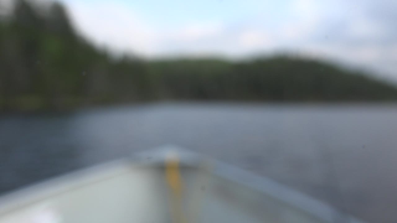 A still-blurry Red Lake, Canada view shows the boat bow centered low in frame, rippled water across the middle, dark green shoreline beyond, and no visible people.