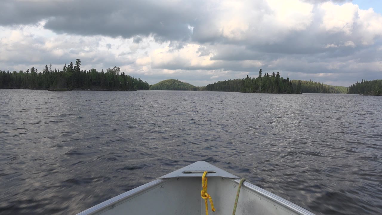 Boat bow on choppy lake facing forested islands under gray clouds.
