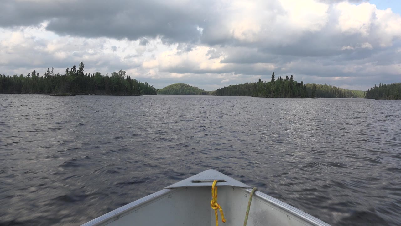 The boat bow remains centered as dark lake ripples lead toward tree-covered islands and a low hill in Red Lake, Canada, with no visible people.