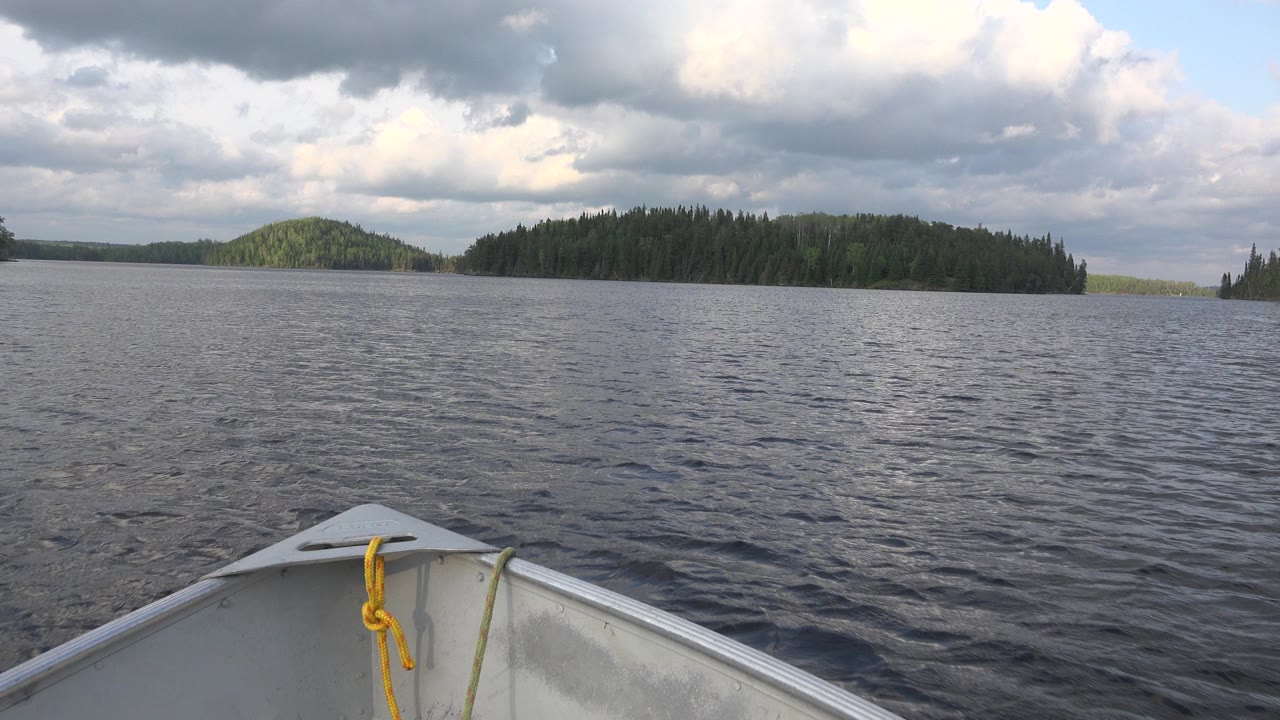 Boat bow on rippled lake facing forested islands under gray clouds.