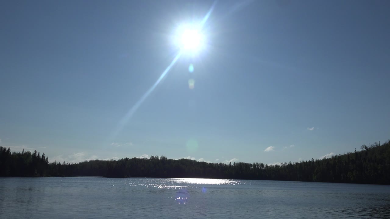 Opening frame at Red Lake Canada with bright sun high over calm lake water, a dark tree-lined shore, blue sky, lens flare, and no visible people.