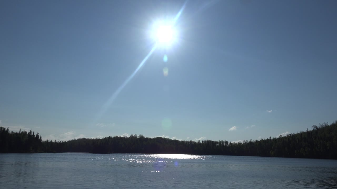 Clear blue sky, high sun, calm water, and wooded shoreline.