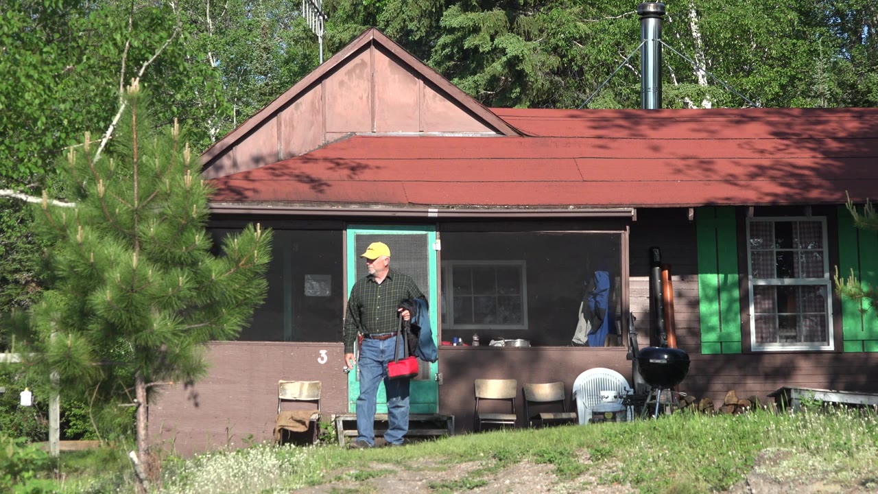 At the start of the Red Lake, Canada clip, an adult man stands near the green-trimmed cabin doorway holding a red bag and a dark jacket.