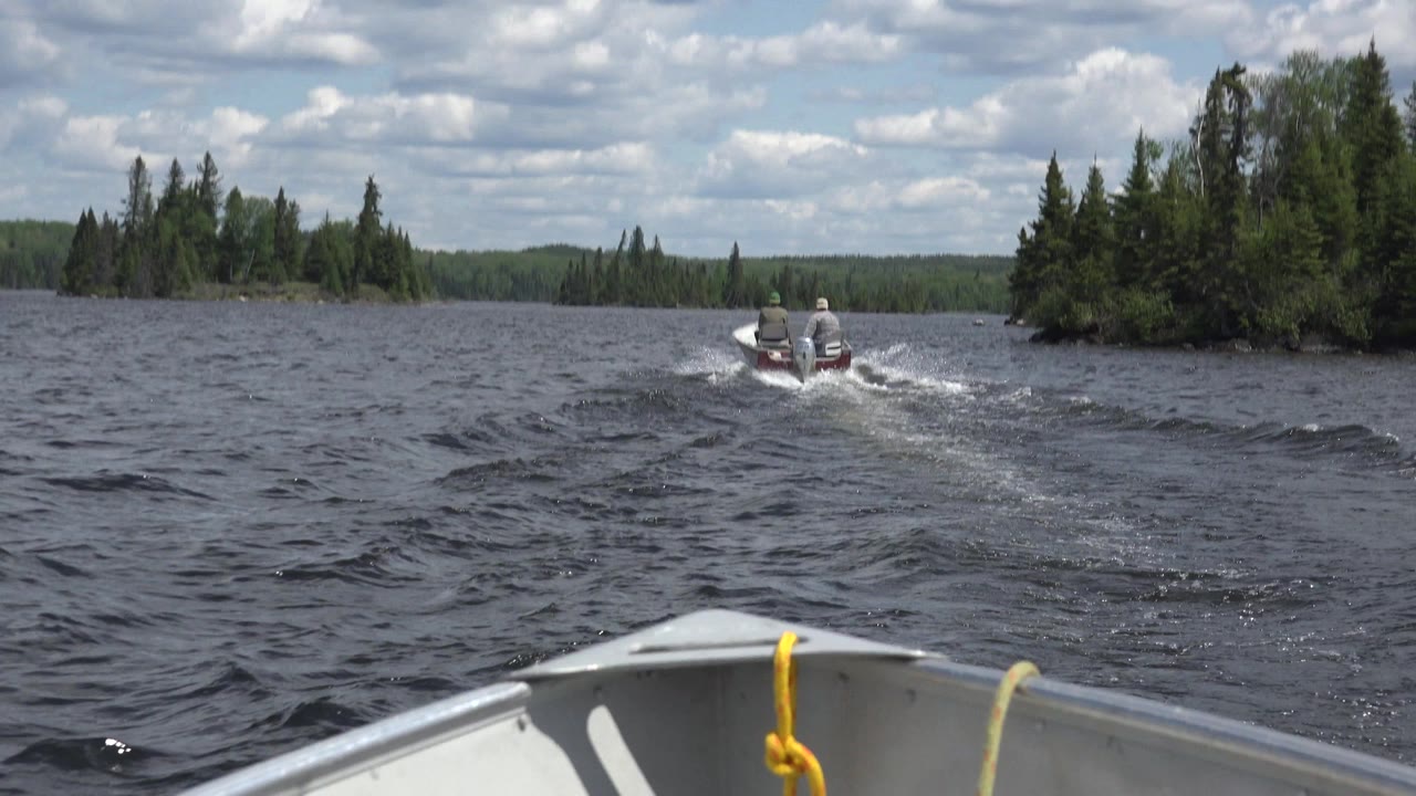 At 0:01 in Red Lake, Canada, the lead motorboat sits farther right on the water near a tree-covered shoreline, leaving a white wake under bright clouds.