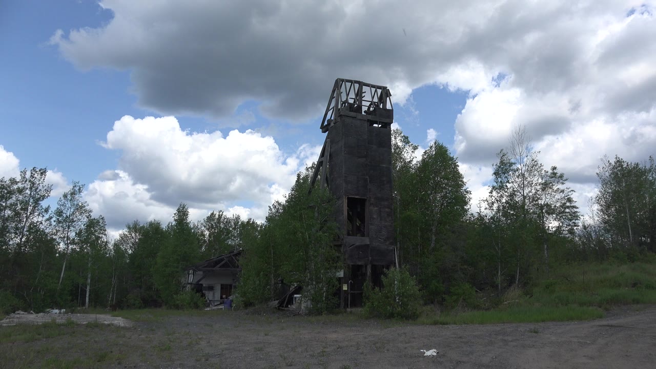 At 0:00 in Red Lake, Canada, a dark wooden tower rises from a gravel clearing with green trees behind it and no visible people.