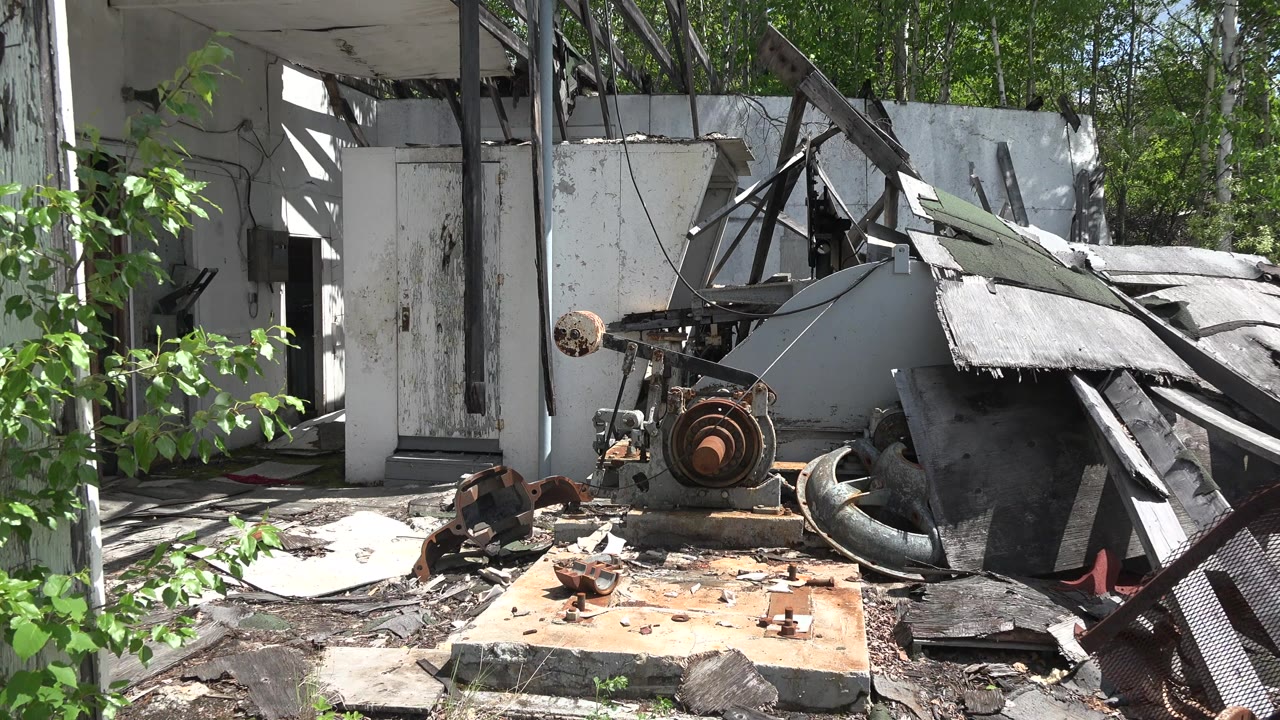 At 0:00 in Red Lake, Canada, a damaged white-walled structure sits beside rusted machinery, broken panels, scattered rubble, and green trees, with no visible people.