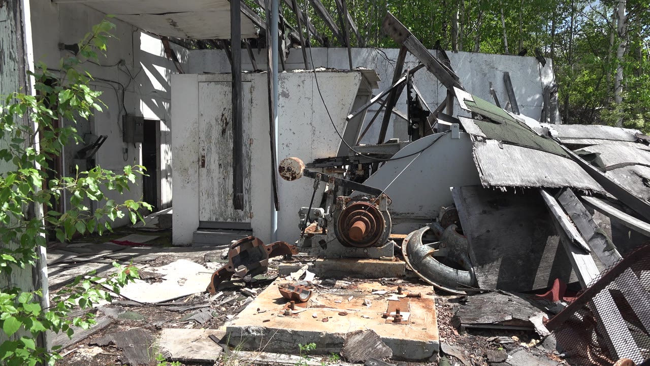 Collapsed roof panels and rusted debris in an overgrown lot.