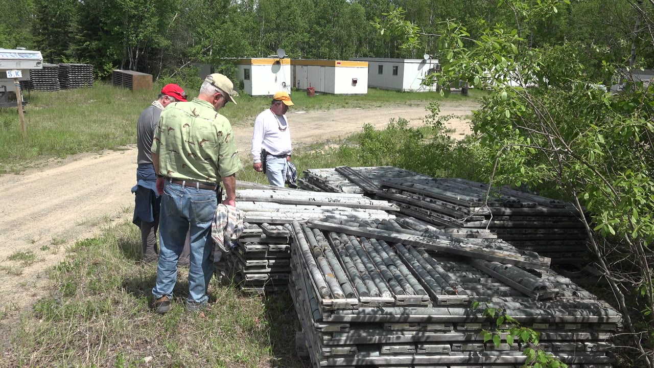 Three adult men in caps stand beside stacked gray metal structures in a grassy yard in Red Lake, Canada, with a dirt road and white trailers behind them.