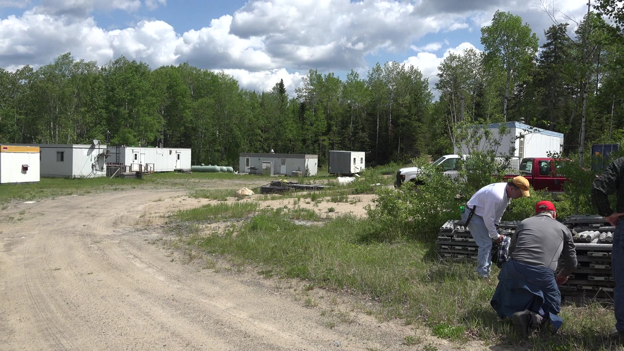 Dirt road beside stacked wooden boards and two men near a rural clearing.