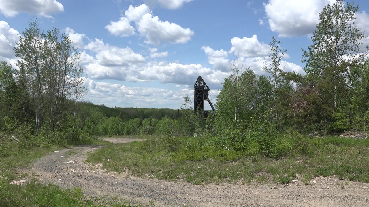 Opening frame of a Red Lake, Canada dirt road curving past green brush toward a tall dark structure near the trees, with no visible people.