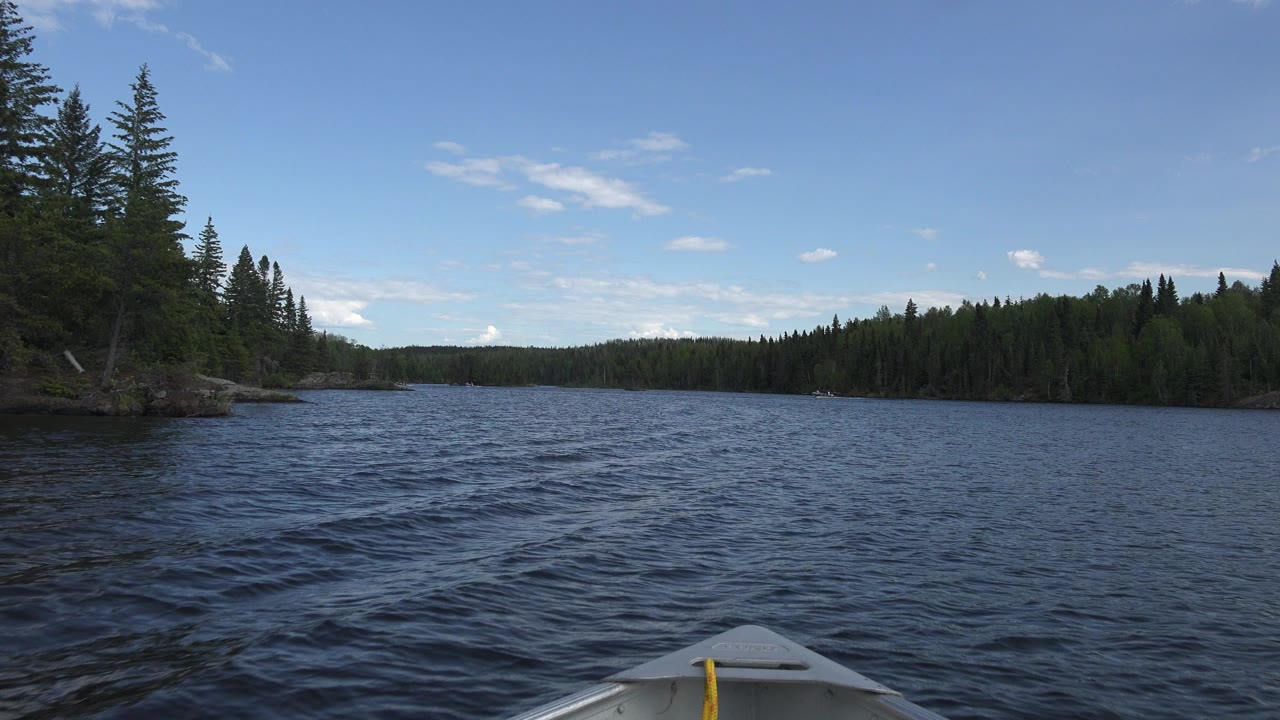Opening frame from Red Lake, Canada shows a small boat bow at the bottom edge, dark rippled water ahead, evergreen trees on both shorelines, scattered clouds, and no visible people.
