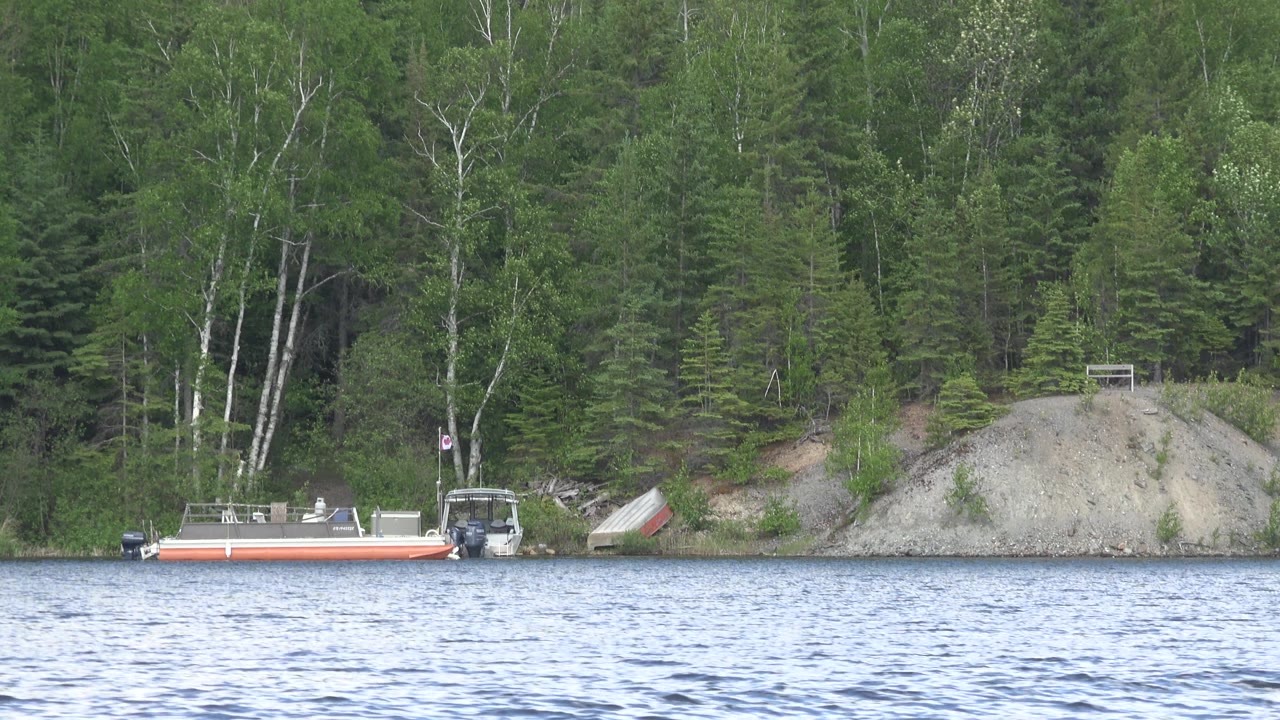 At Red Lake, Canada, the same shoreline boat remains near the left of frame while the pale rock mound and dense green trees sit behind it, with no visible people.