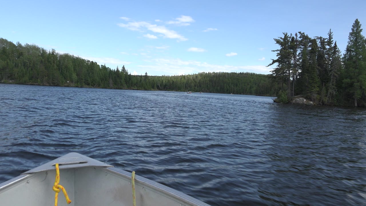 At 0:00 in Red Lake, Canada, a small aluminum boat bow with a yellow rope points toward rippled blue water, a tree-lined shore, and no clearly visible people.