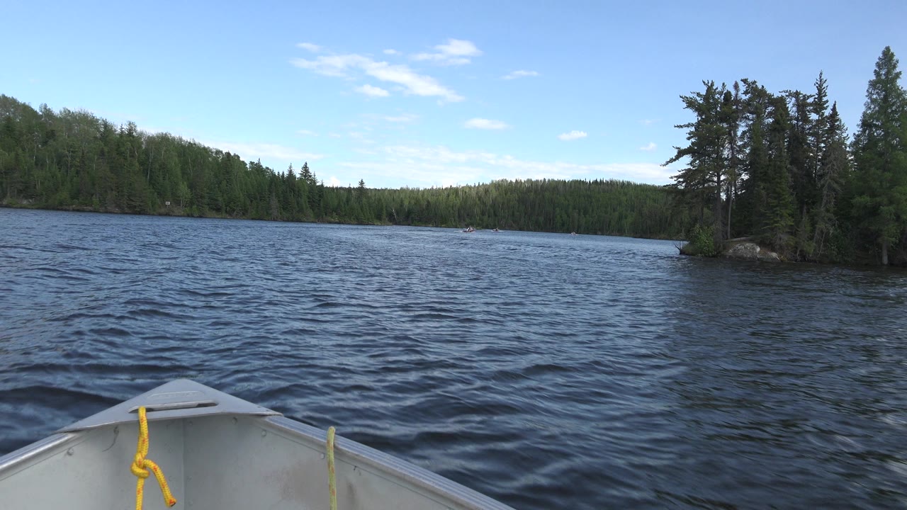 At 0:01 in Red Lake, Canada, the boat bow remains in the lower frame as darker ripples lead toward a small tree-covered point and distant wooded bank, with no clearly visible people.