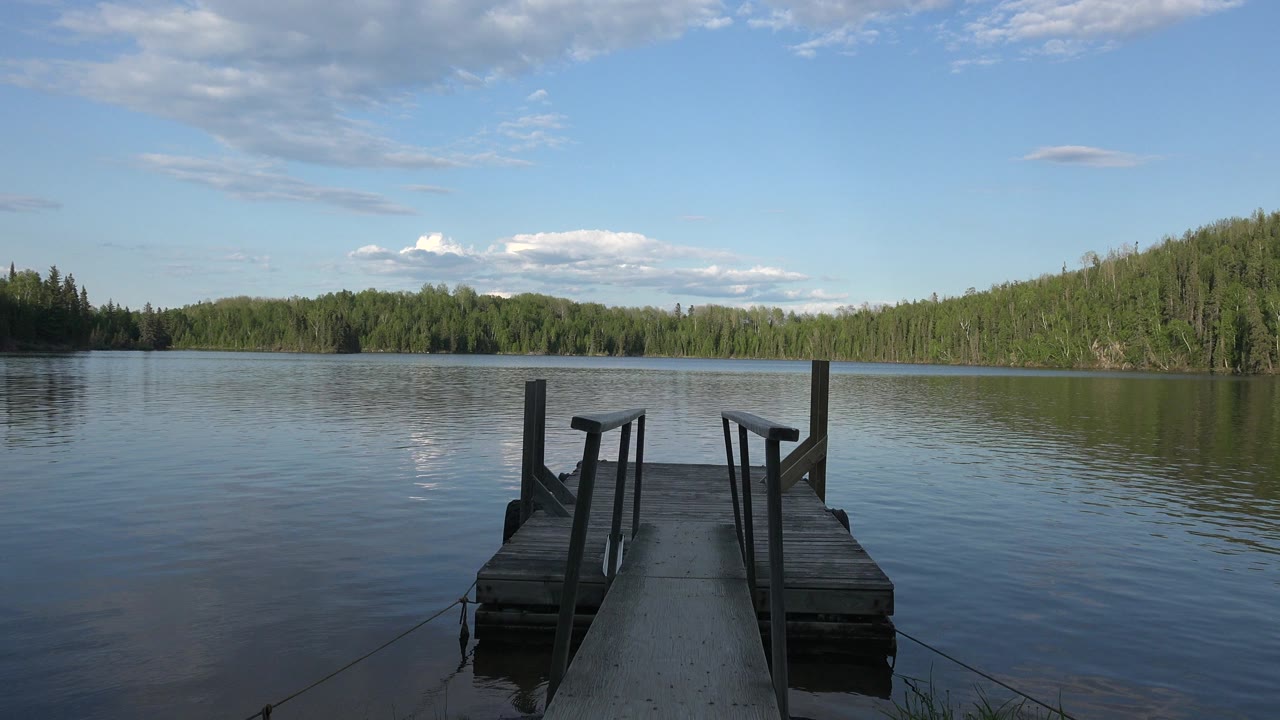 Opening frame of a wooden pier at Red Lake, Canada, extending into calm water toward a green tree-lined shore under blue sky, with no visible people.