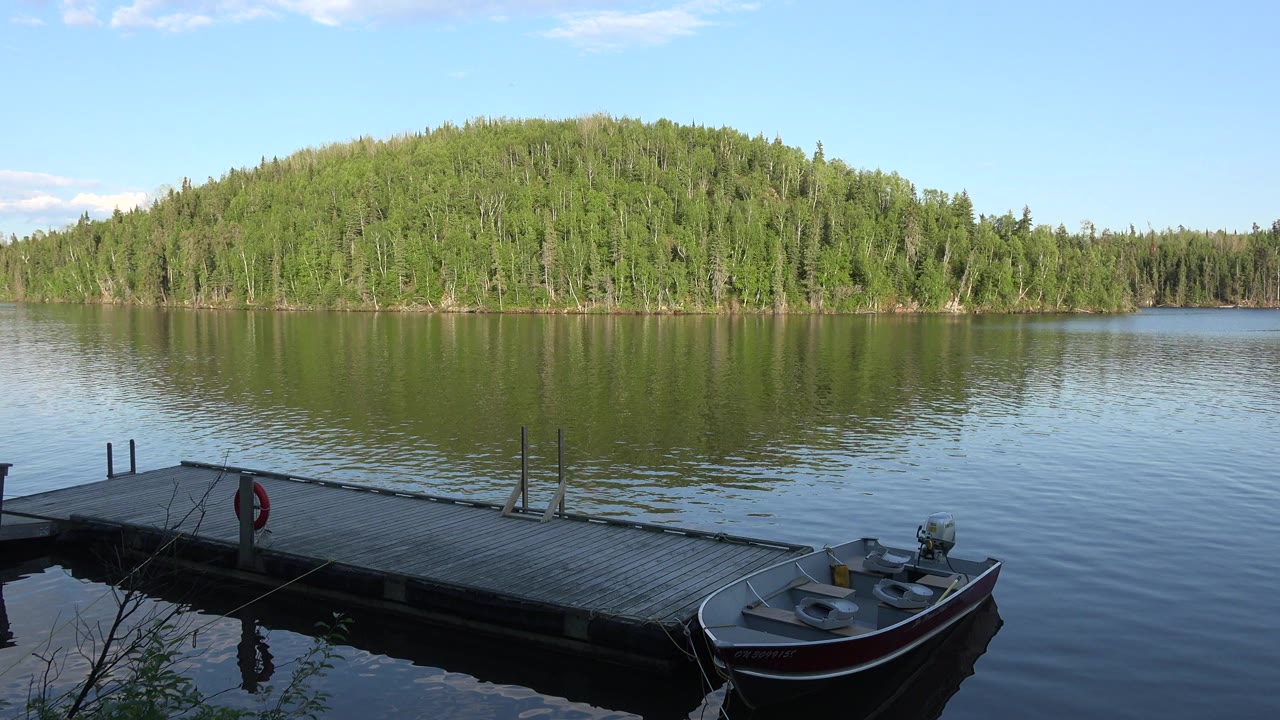 Opening frame in Red Lake, Canada showing a small boat tied near a wooden dock, calm water, a tree-covered island, pale blue sky, and no visible people.
