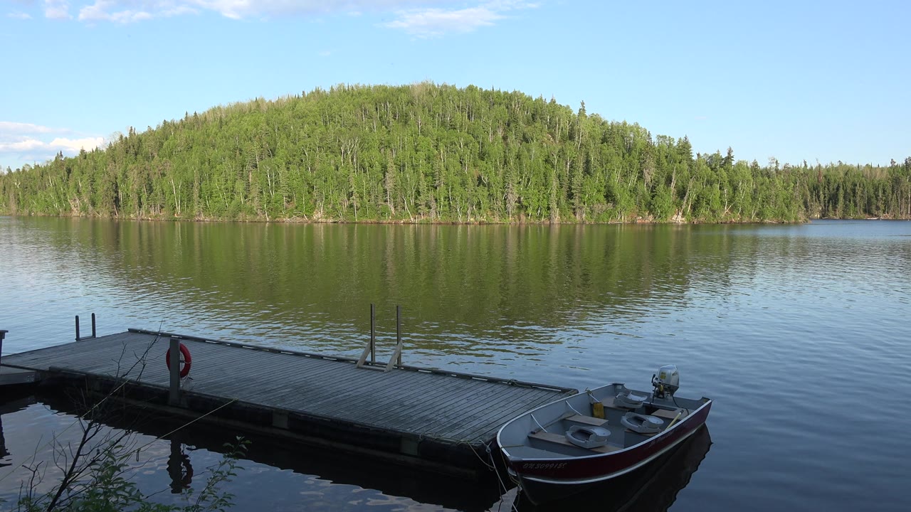 Boat moored beside a dock with calm lake water and wooded shoreline in the distance.