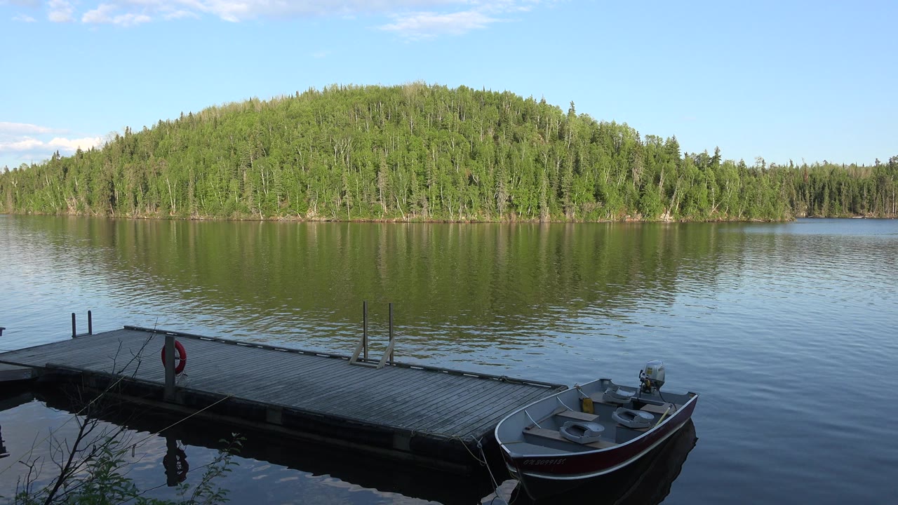 Opening frame of a Red Lake, Canada dock with a small fishing boat in the shaded foreground, calm water, a green forested hill, and no visible people.