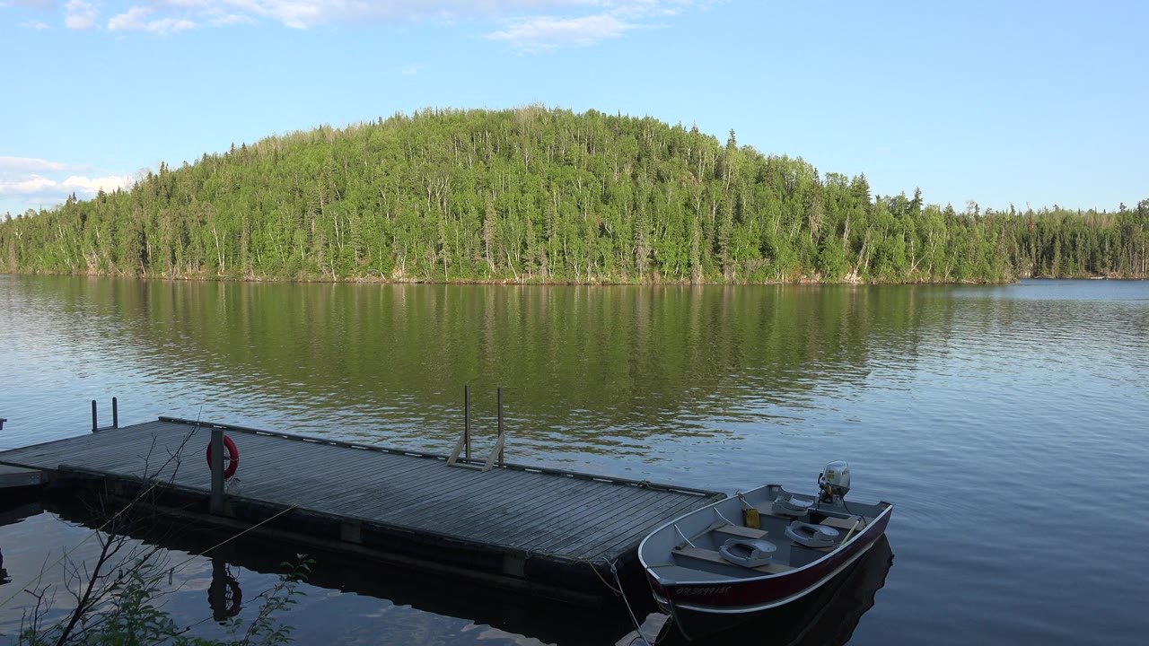 At Red Lake Canada, a dock and small motorboat sit in the foreground while calm water reflects a tree-covered hill, with no visible people.